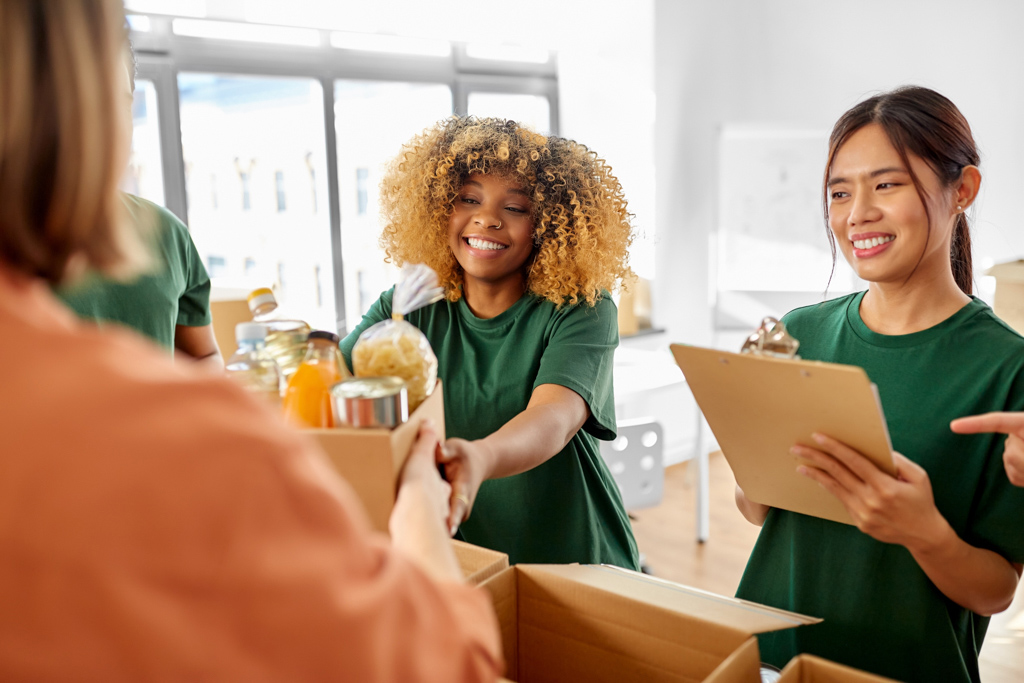 One adult female smiling while handing a bag of groceries over to an adult female, an adult female smiling while holding a clipboard.