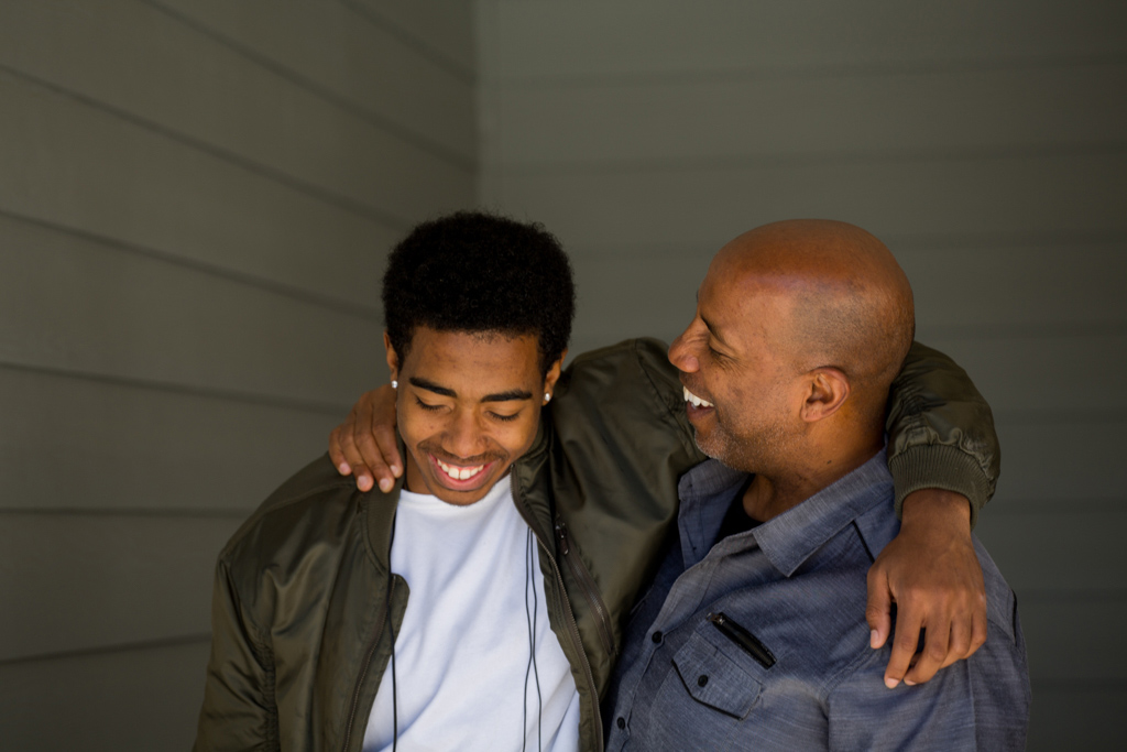 Male and teenage boy standing side by side with arms around each other's shoulders, smiling.