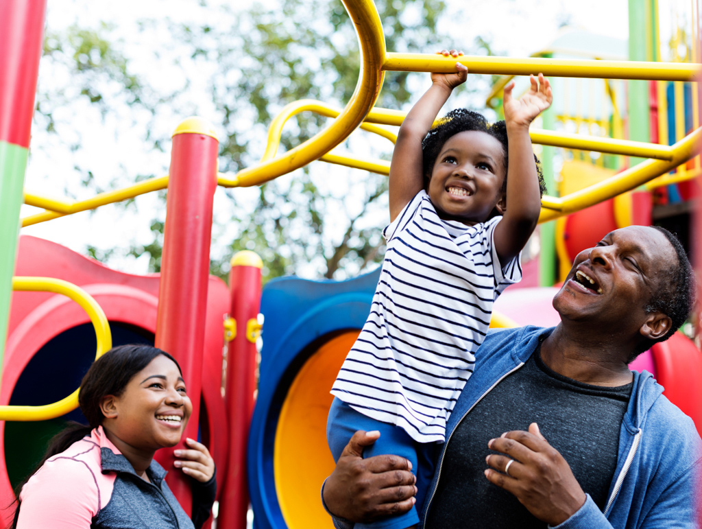 Older male holding little girl while she plays on the monkey bars, while a woman looks on smiling. 