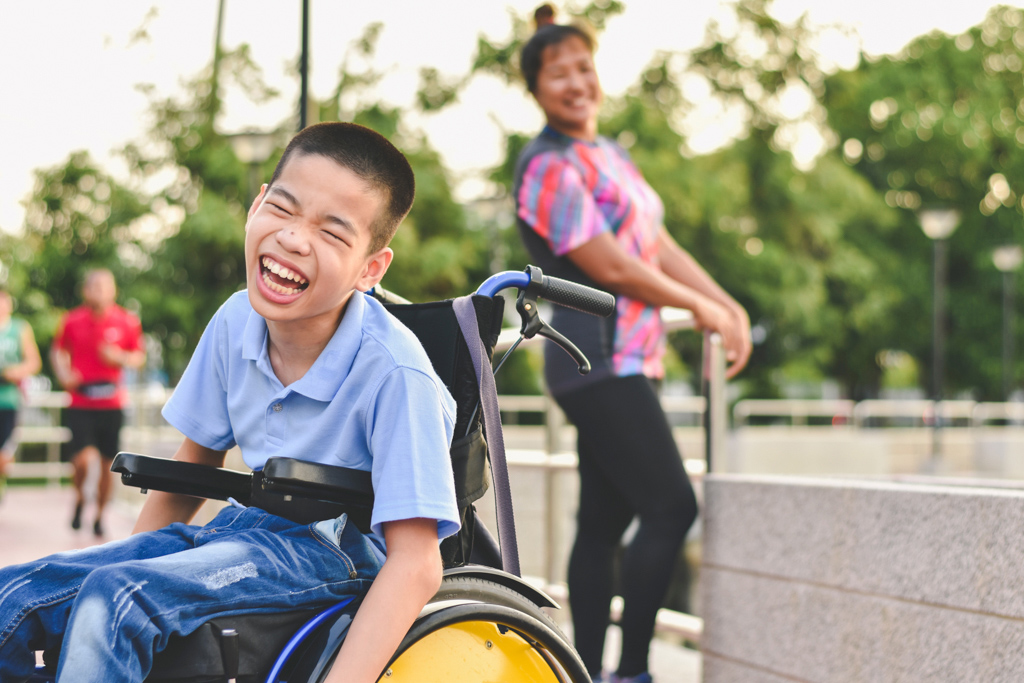 A young boy sitting in a wheelchair outside smiling.