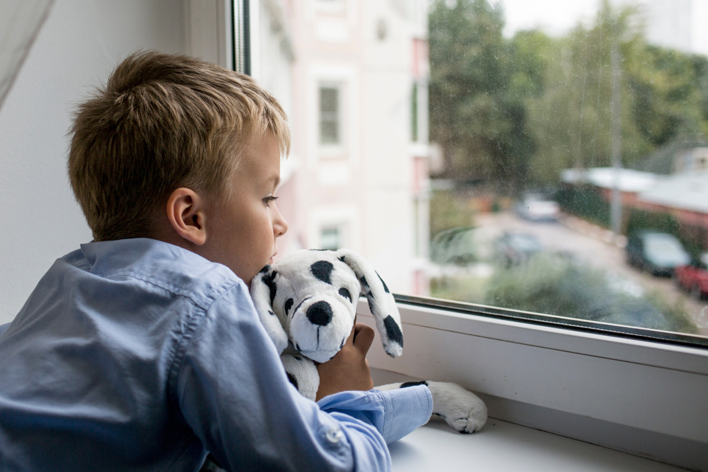 Young boy looking out window while holding stuffed animal.