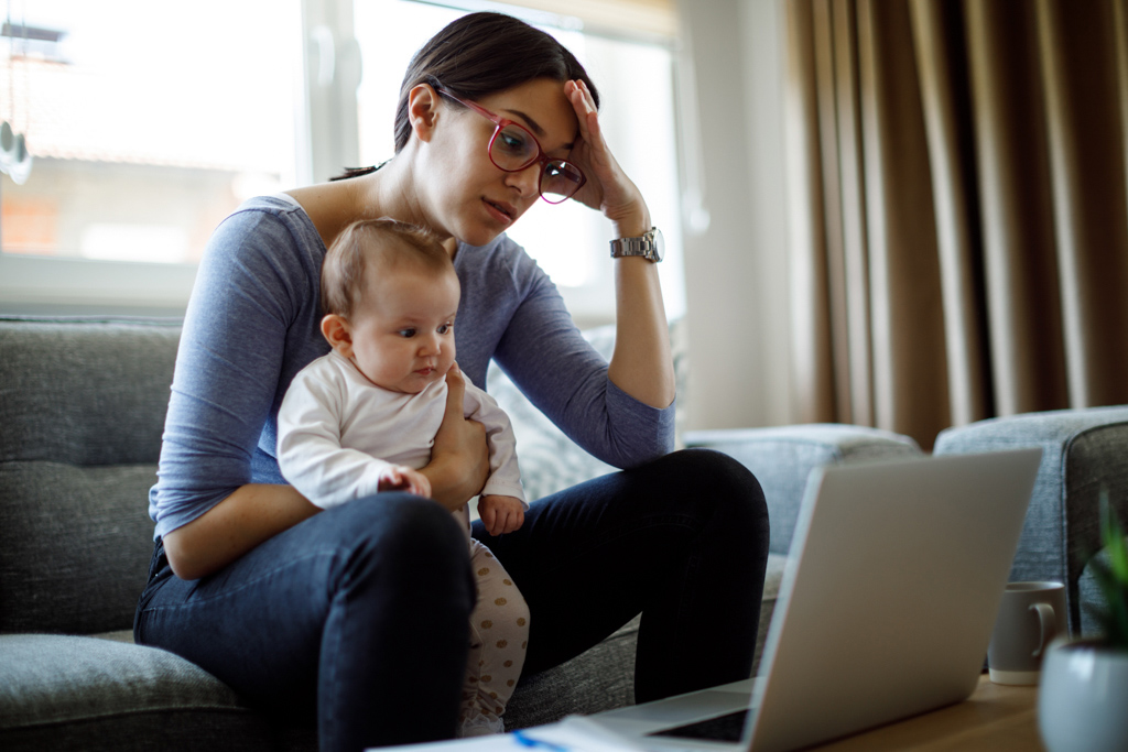 Woman holding baby on lap while sitting on the couch, staring at laptop with perplexed face.