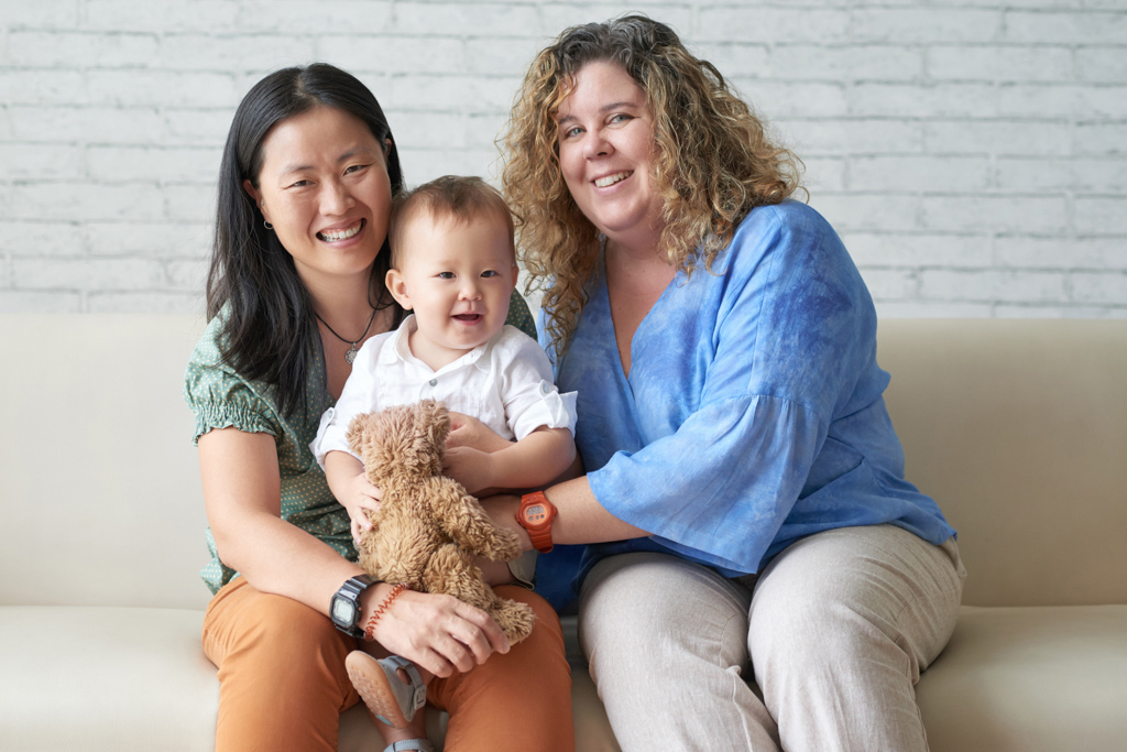 Two women holding baby while sitting on sofa, smiling.