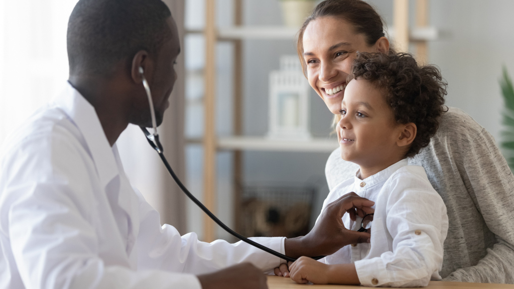 An adult male putting a stethoscope on a young child who is sitting in the lap of an adult woman.