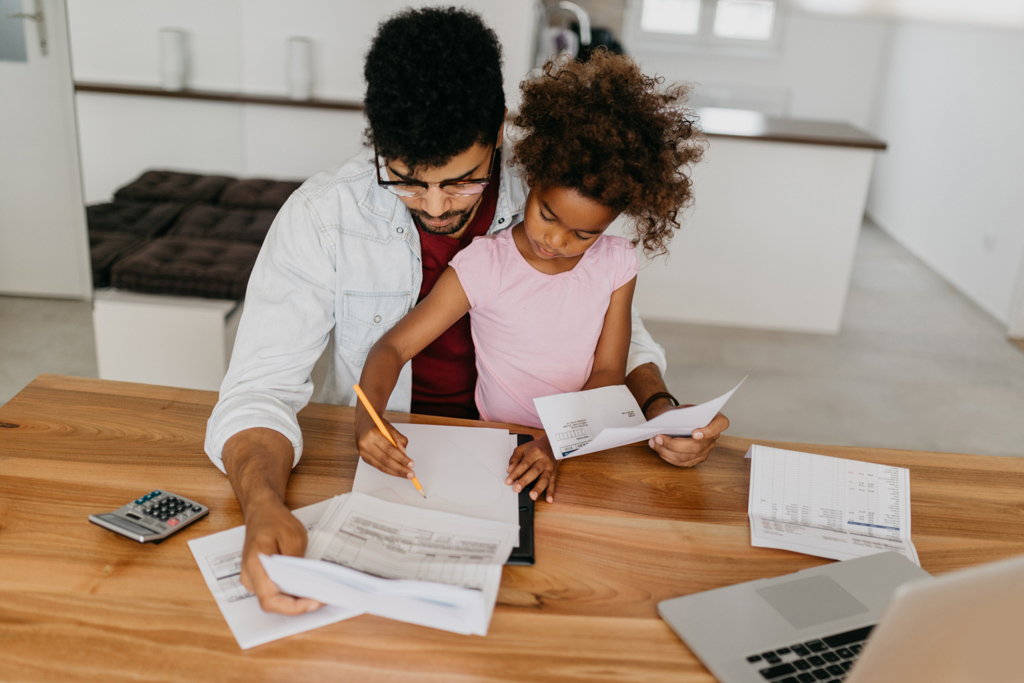 Man sitting at table filled with papers, computer, and a calculator, with young girl sitting on his lap, while they both stare at papers.