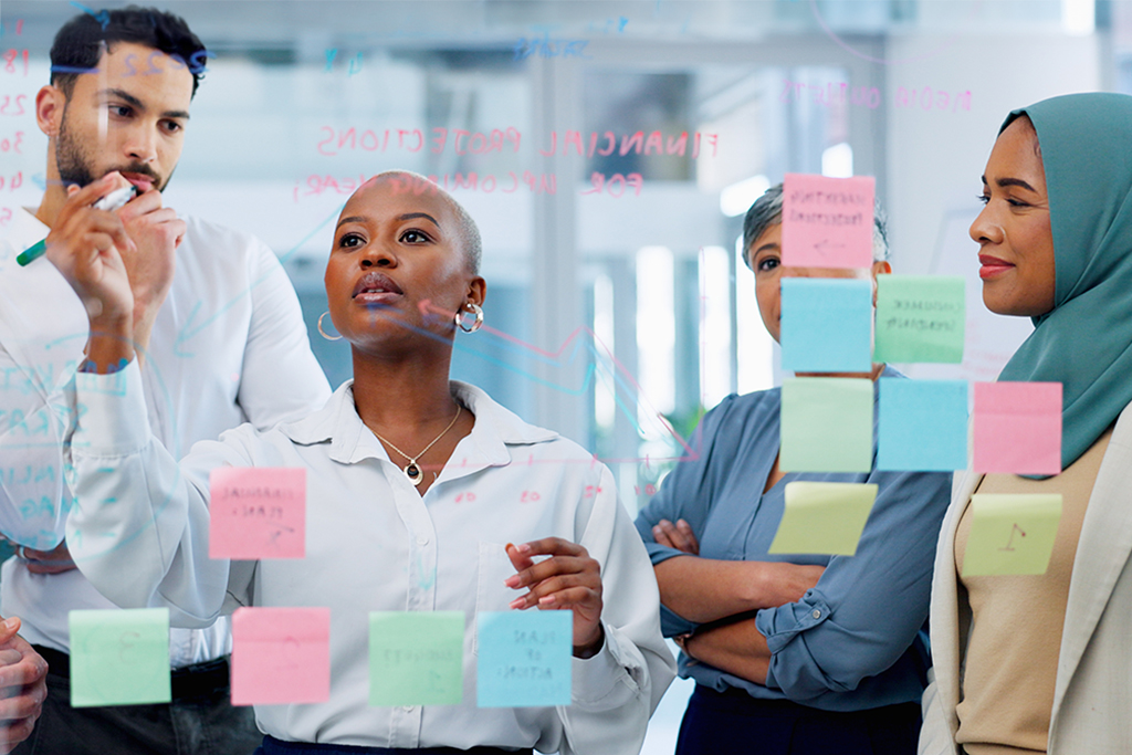 A man and three women dressed in business casual clothing standing together as one woman writes notes on a glass wall filled with sticky notes.