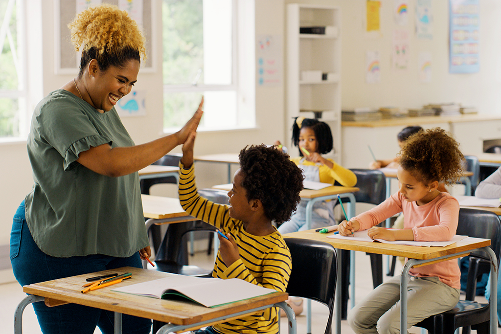 A classroom scene with a teacher and three students seated at desks. The teacher, standing, is giving one student a high five.