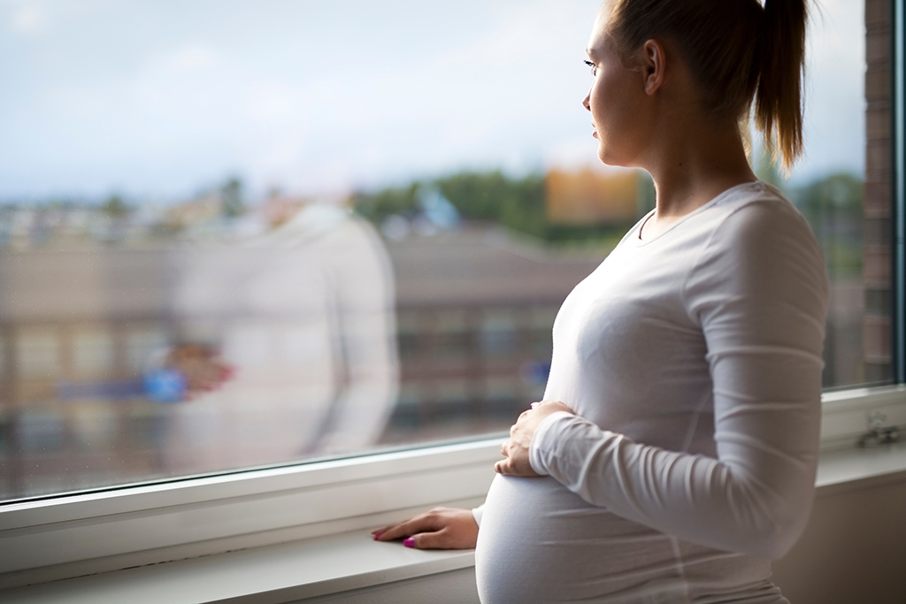 Young pregnant woman looking out the window with hand on her stomach.