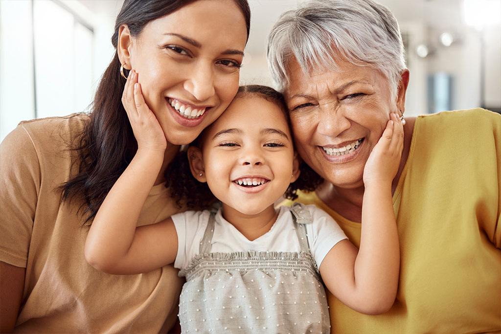 a female child sitting with her hands raised up and placed on the cheeks between an adult female and a female grandparent