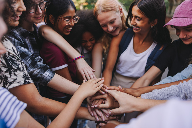 Diverse group of young people standing in a circle with their hands placed in the middle on top of each other.