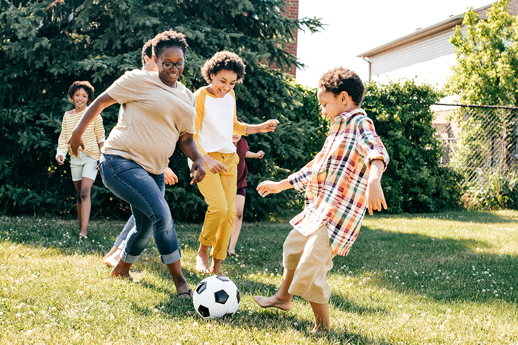 A group of adults and children playing soccer in a backyard on a sunny day.