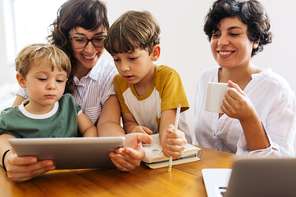 Two women and two young boys sitting together at table looking at tablet, smiling.