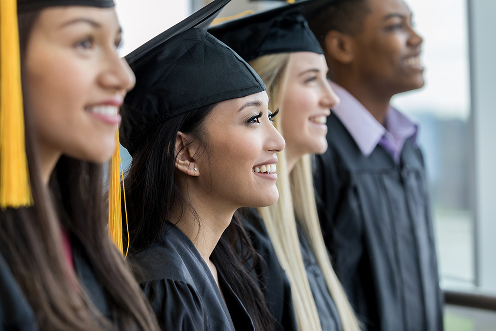 hree high school girls and one boy standing in a row wearing a cap and gown for  graduation, smiling. 