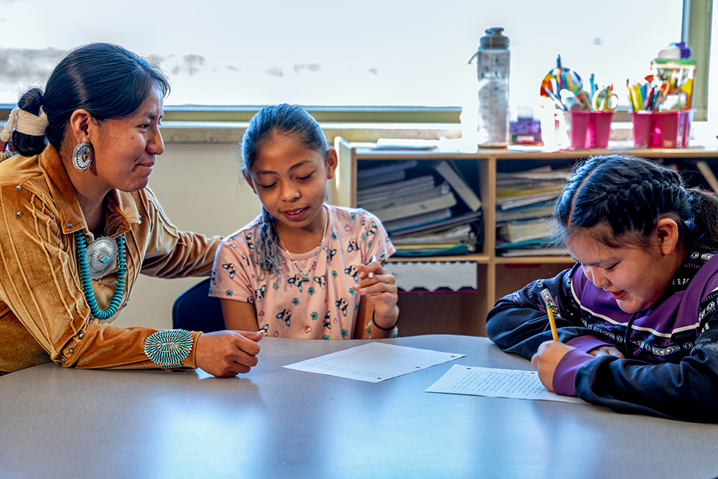 An indigenous woman assisting elementary age students in a classroom as they sit at a table together.