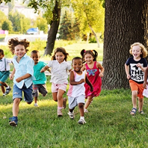A diverse group of young children happily running outside through a tree filled park on a sunny day.