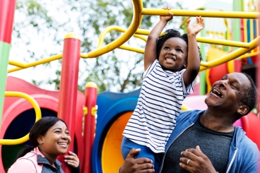 Older male holding little girl while she plays on the monkey bars, while a woman looks on smiling. 