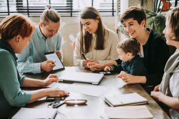 Young woman giving a presentation with tablet at conference table with four other women and one child.