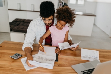 Man sitting at table filled with papers, computer, and a calculator, with young girl sitting on his lap, while they both stare at papers.