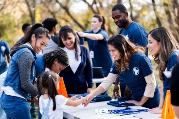 An adult woman reaches across the table to shake hands with a young girl while surrounded by a group of adults wearing matching t-shirts.