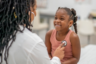 A healthcare professional is using a stethoscope to listen to the chest of a young girl sitting on an examination table