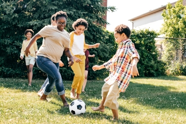 A group of adults and children playing soccer in a backyard on a sunny day.