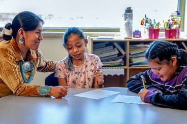 An indigenous woman assisting elementary age students in a classroom as they sit at a table together.