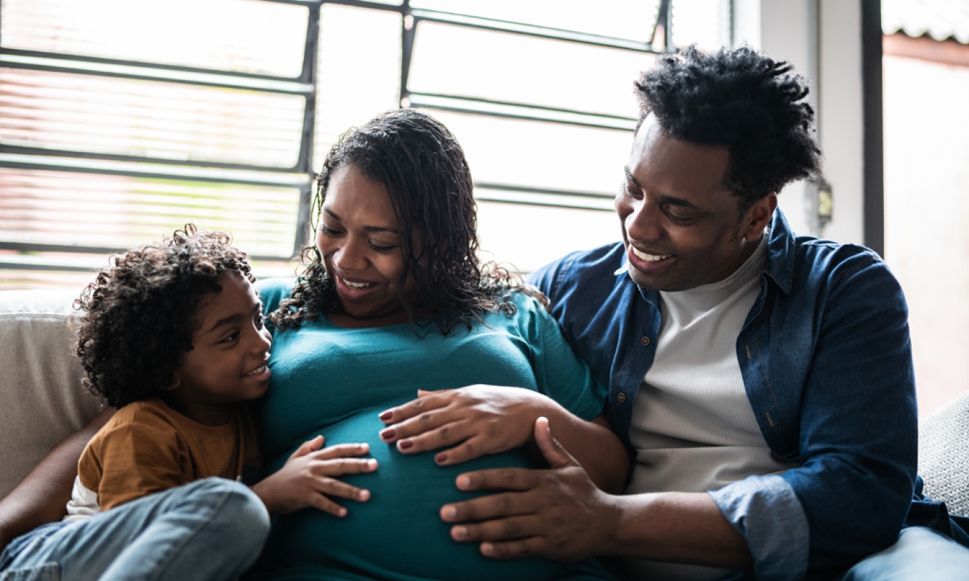 A woman who is pregnant touching her belly, sitting next to a man and child, smiling.