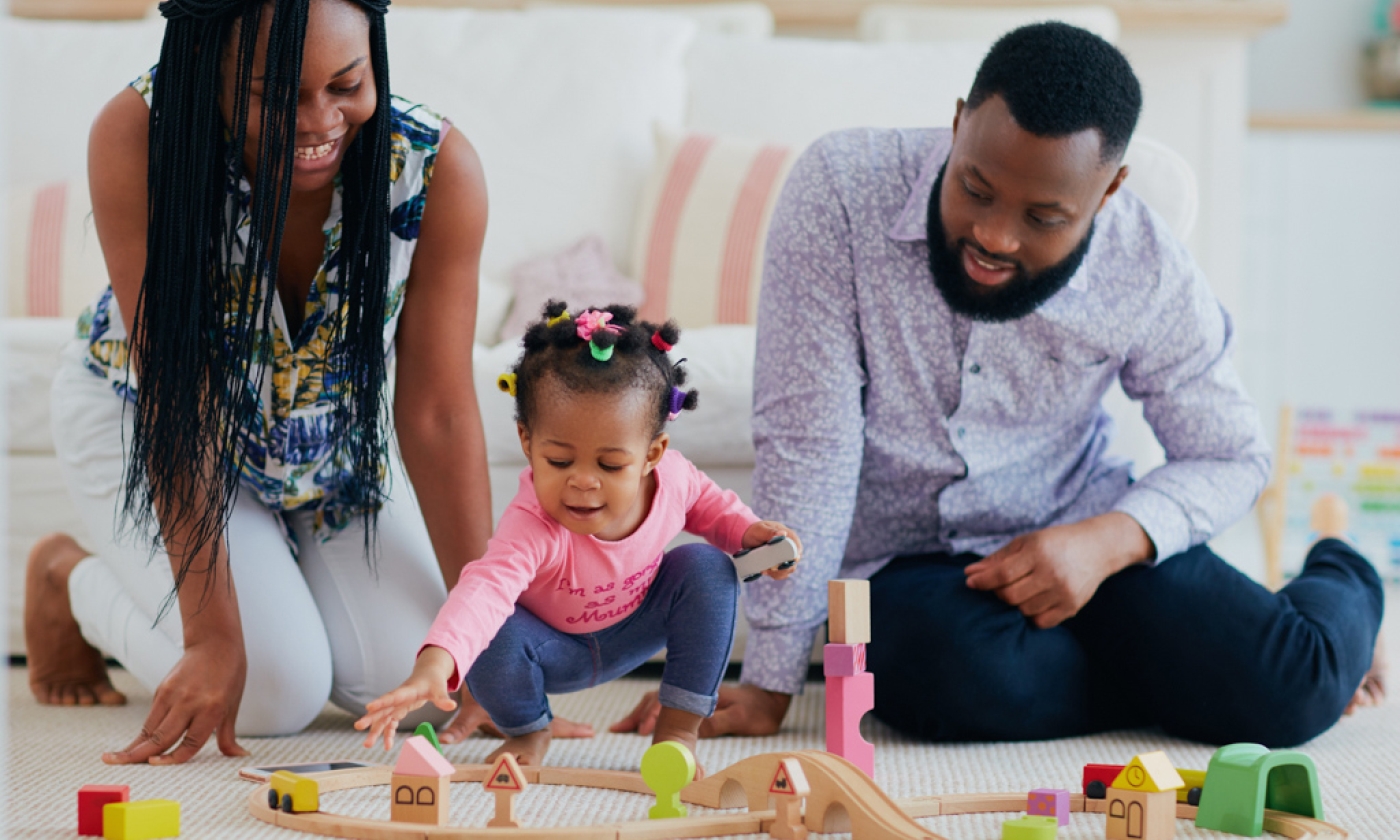 Adult woman and man sitting no floor with little girl between them playing with wooden toys.