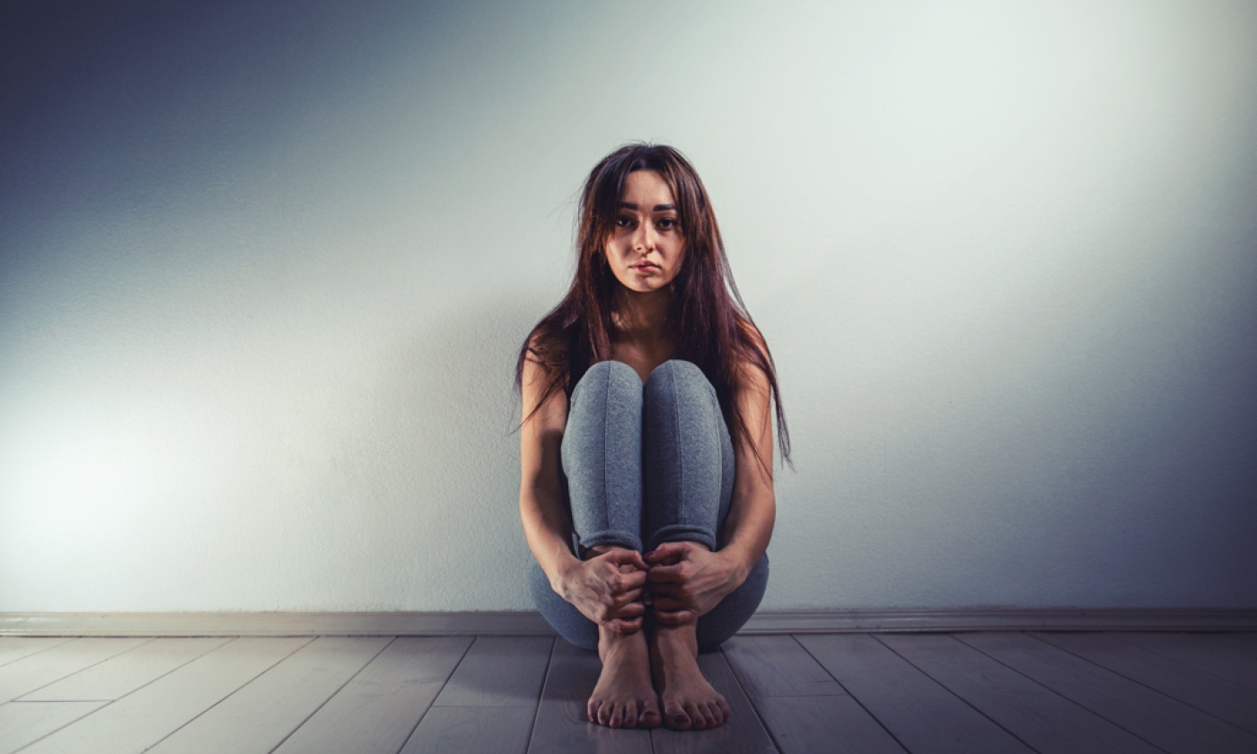A young girl sitting on the floor alone looking alone with her hands around her ankles.