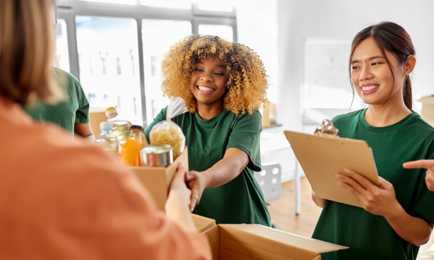 One adult female smiling while handing a bag of groceries over to an adult female, an adult female smiling while holding a clipboard.