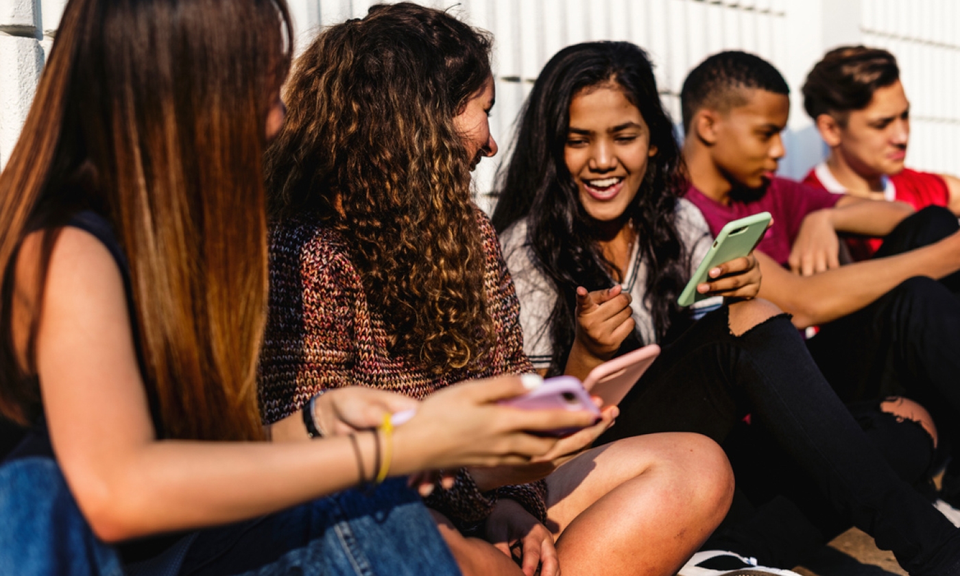 Five young adults sitting against a fence, one of the female young adults is laughing while holding her phone.