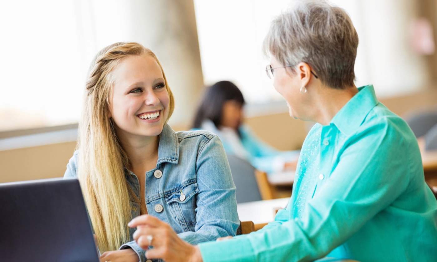 A woman and teen girl sitting at a table in front of a laptop, having a discussion and smiling.