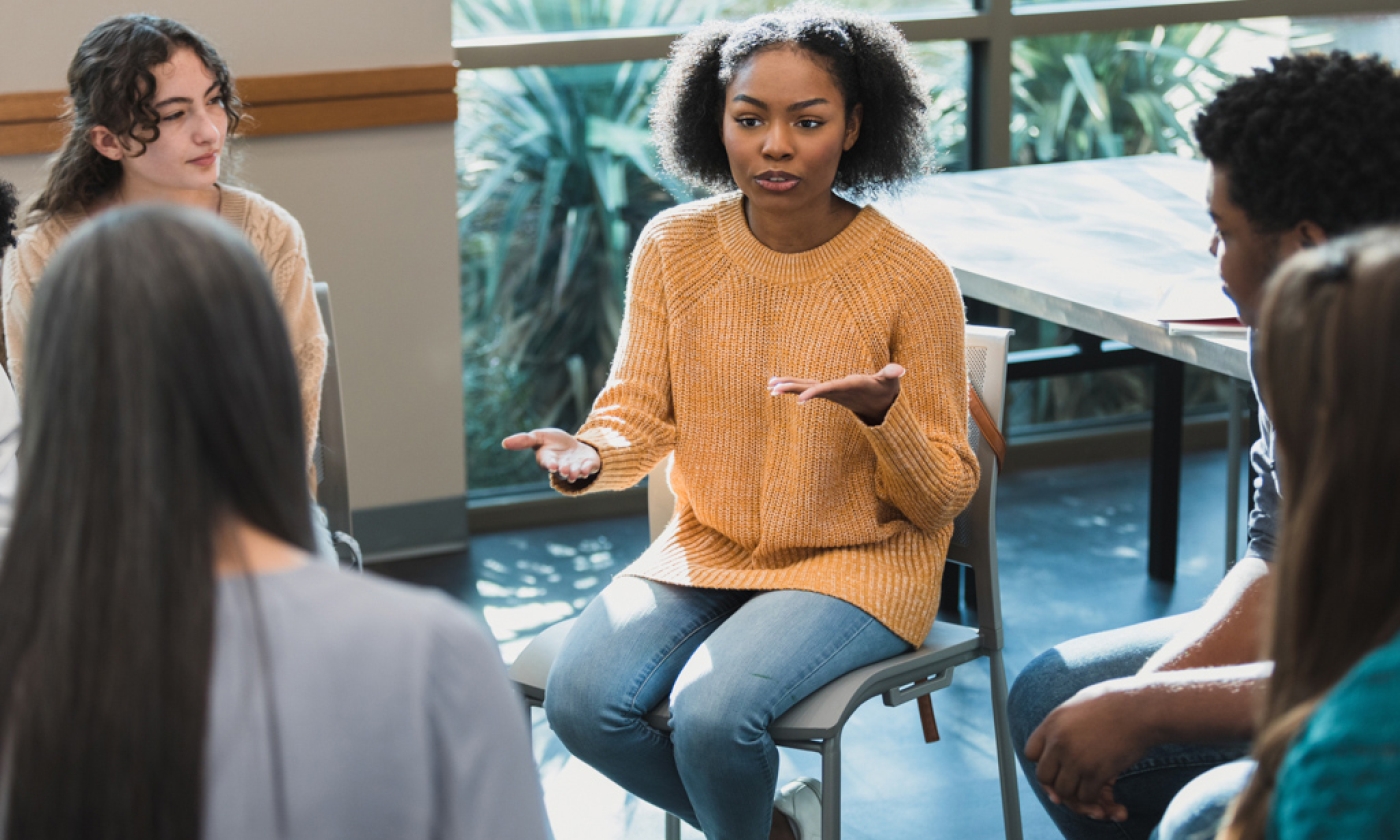 A diverse group of five young adults sit in a circle and focus on one girl who is talking.