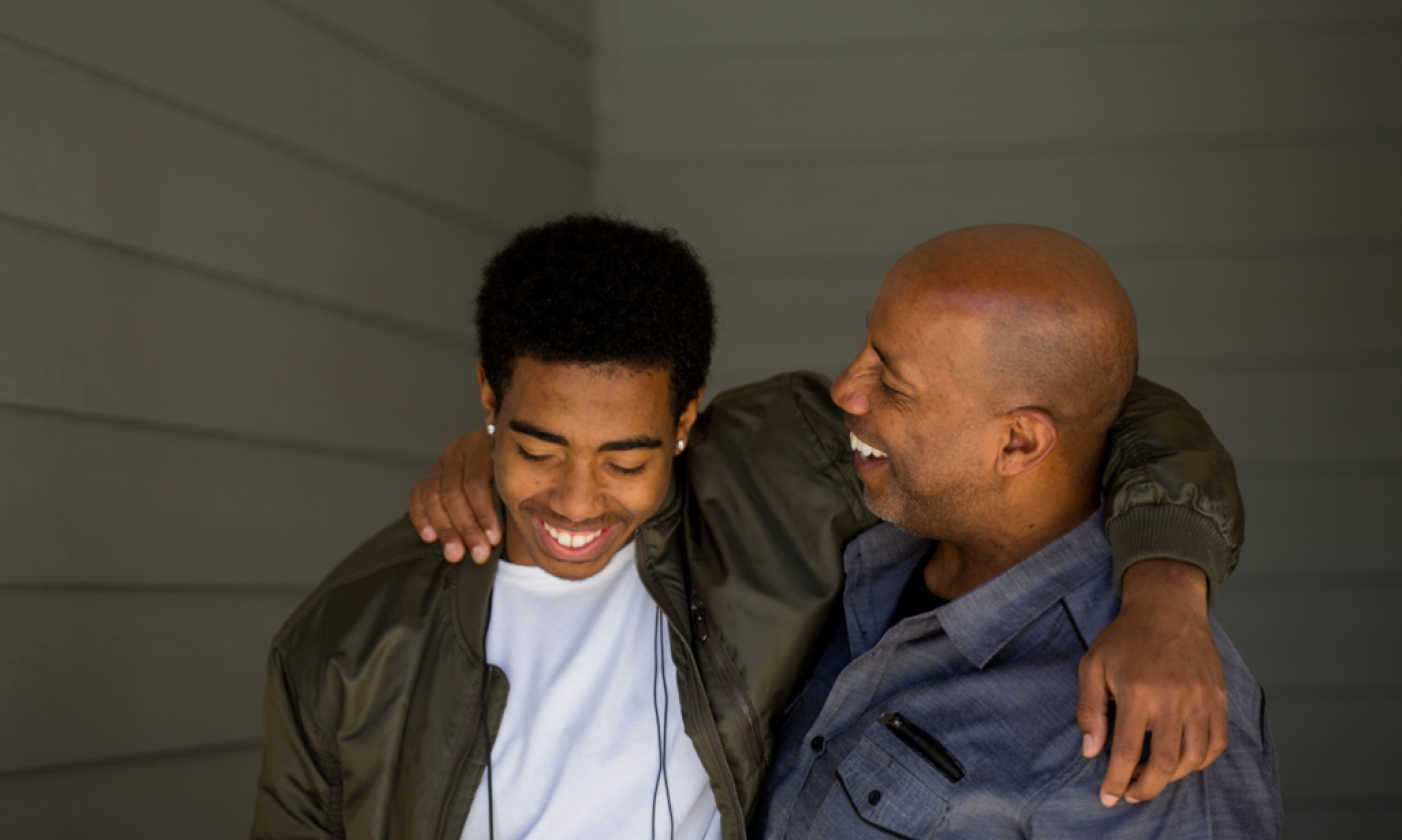 Male and teenage boy standing side by side with arms around each other's shoulders, smiling.