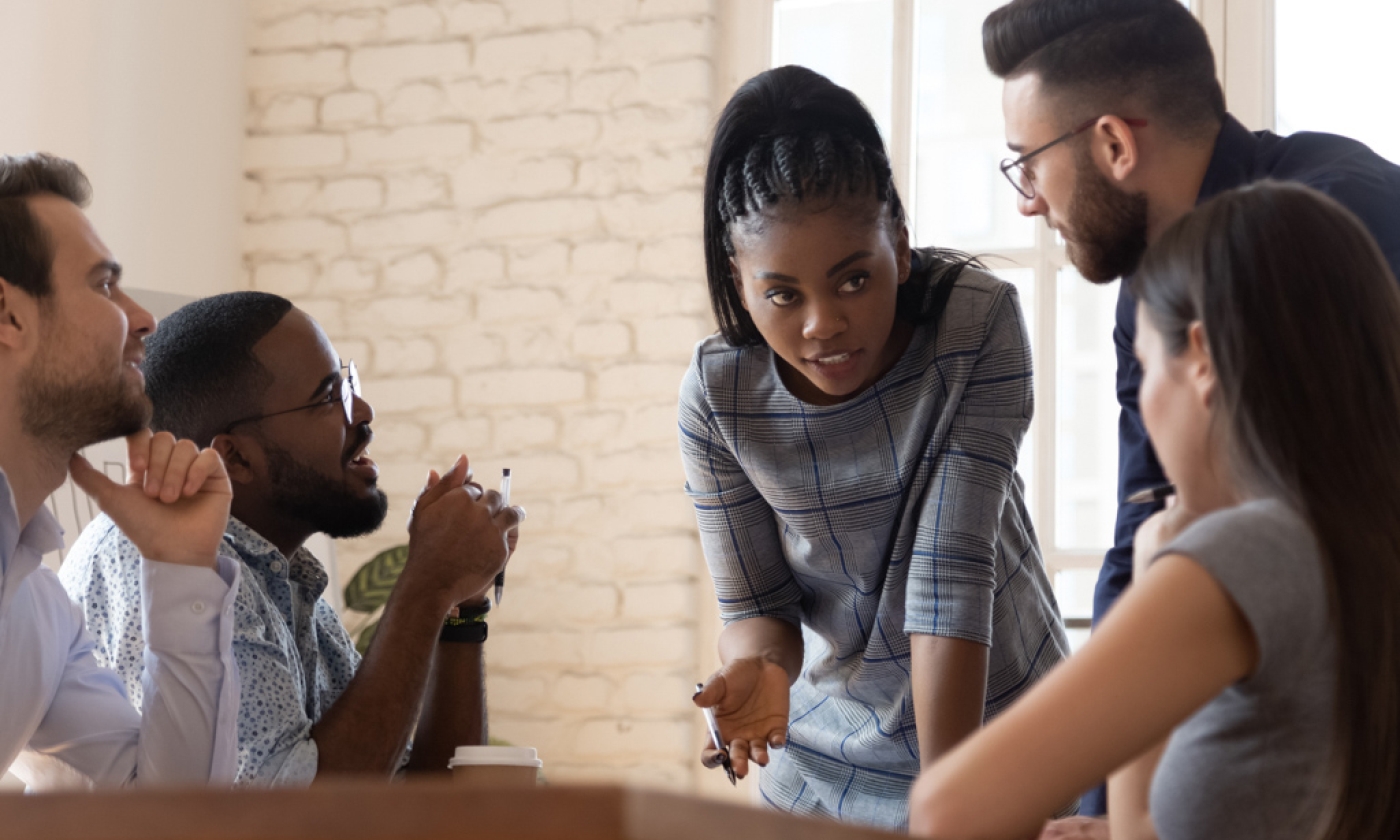 Female employee leaning over conference table while speaking to four coworkers.