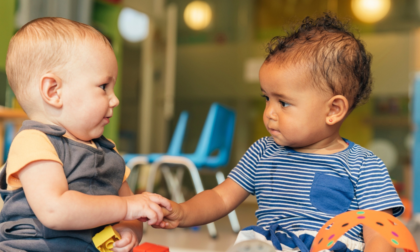 Two babies looking and each other and holding hands while playing with blocks.