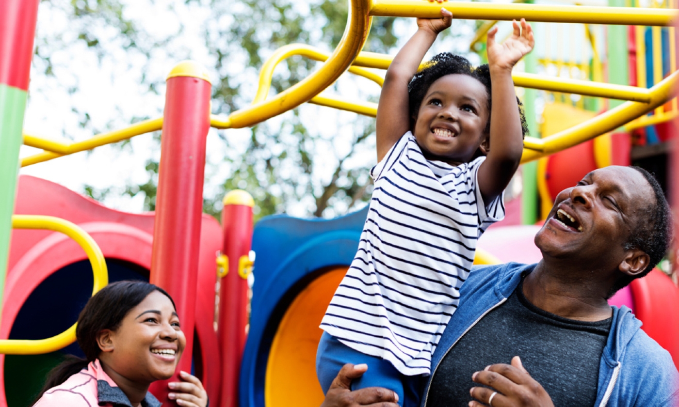 Older male holding little girl while she plays on the monkey bars, while a woman looks on smiling.