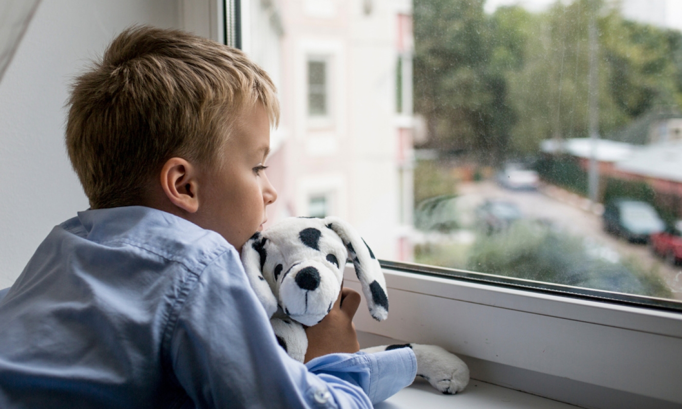 Young boy looking out window while holding stuffed animal.