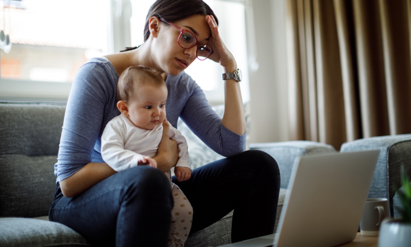 Woman holding baby on lap while sitting on the couch, staring at laptop with perplexed face.