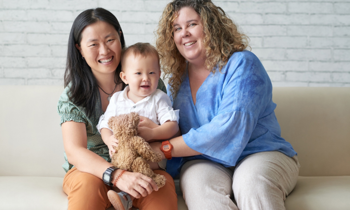 Two women holding baby while sitting on sofa, smiling.