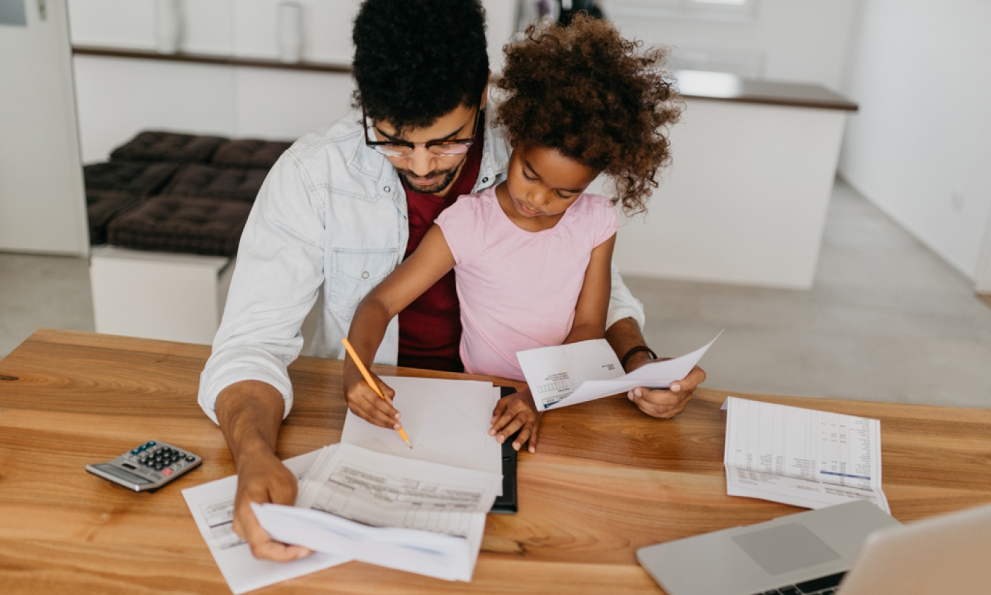 Man sitting at table filled with papers, computer, and a calculator, with young girl sitting on his lap, while they both stare at papers.