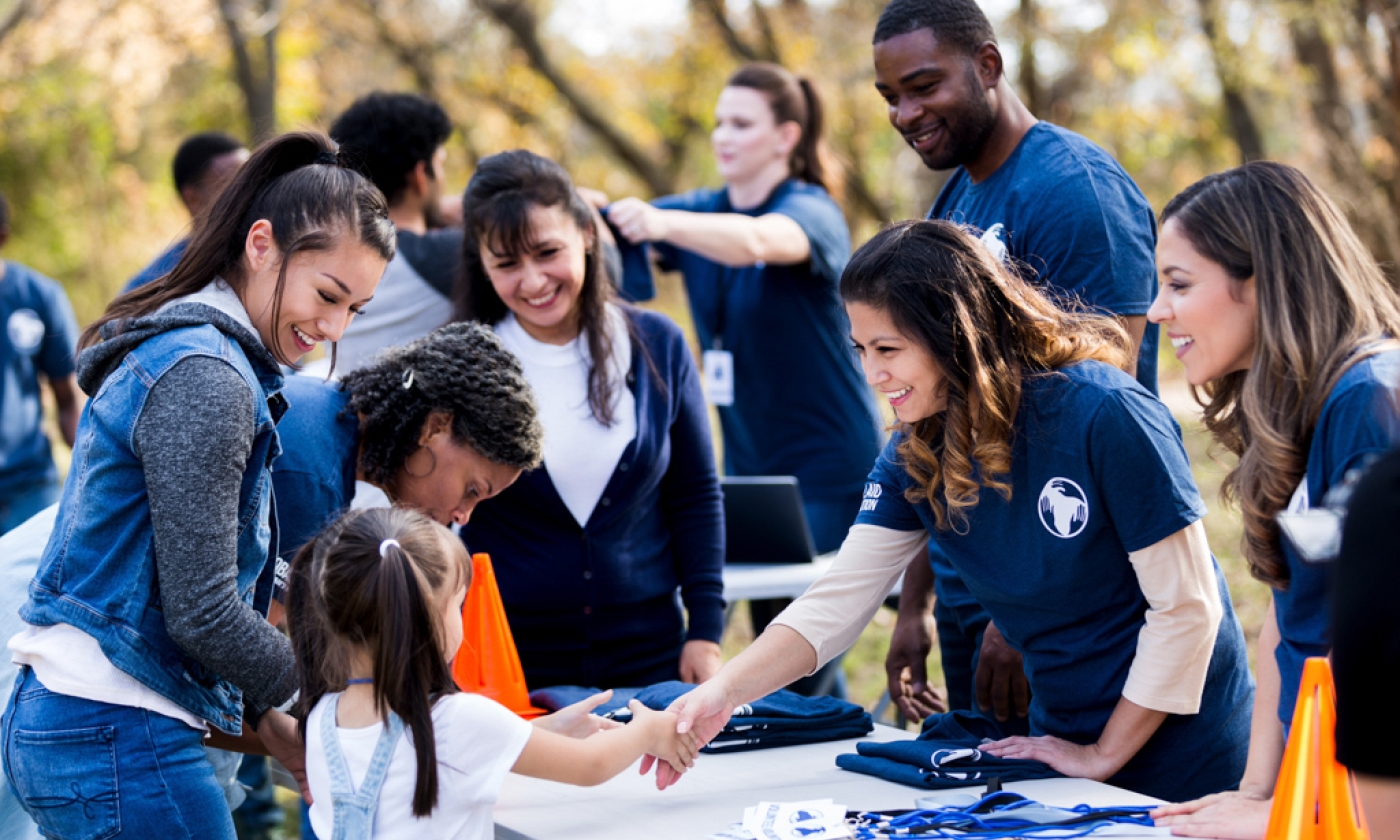 An adult woman reaches across the table to shake hands with a young girl while surrounded by a group of adults wearing matching t-shirts.