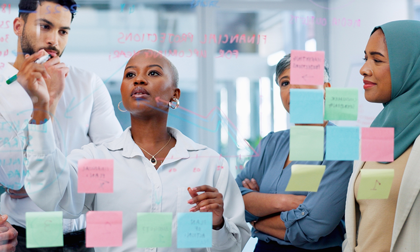 A man and three women dressed in business casual clothing standing together as one woman writes notes on a glass wall filled with sticky notes.
