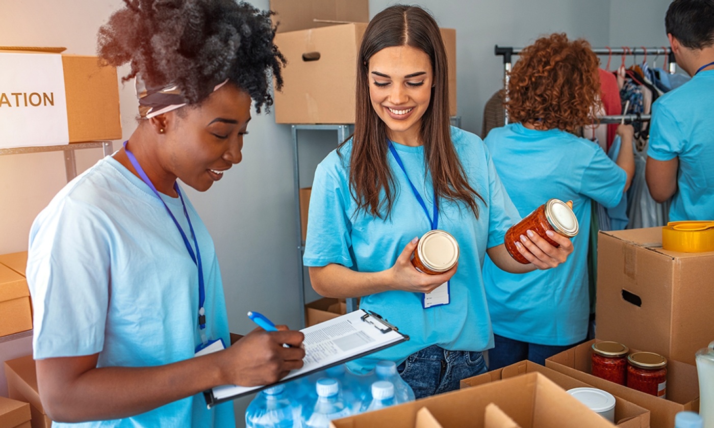 two woman standing next to each other, one holding two canned goods looking down at the clip board the other woman is writing on