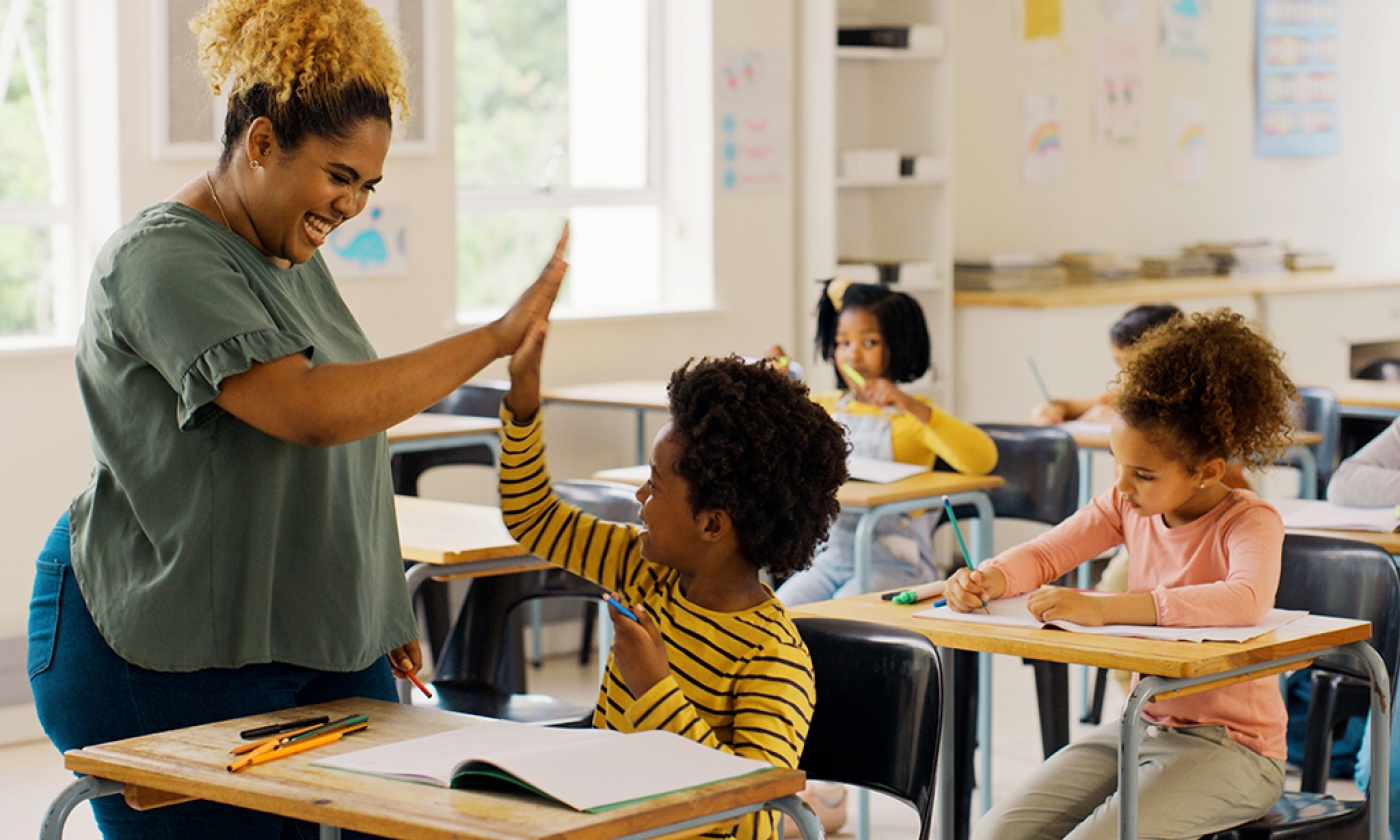 A classroom scene with a teacher and three students seated at desks. The teacher, standing, is giving one student a high five.