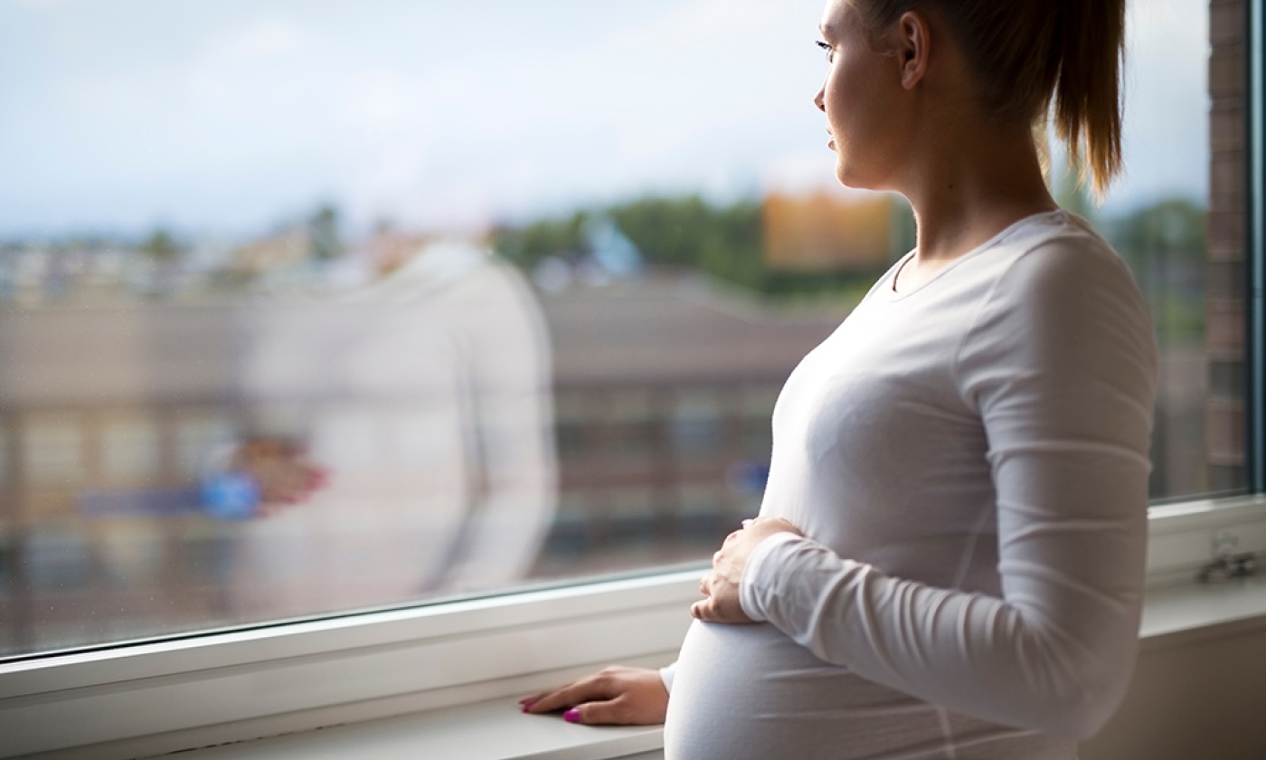 Young pregnant woman looking out the window with hand on her stomach.