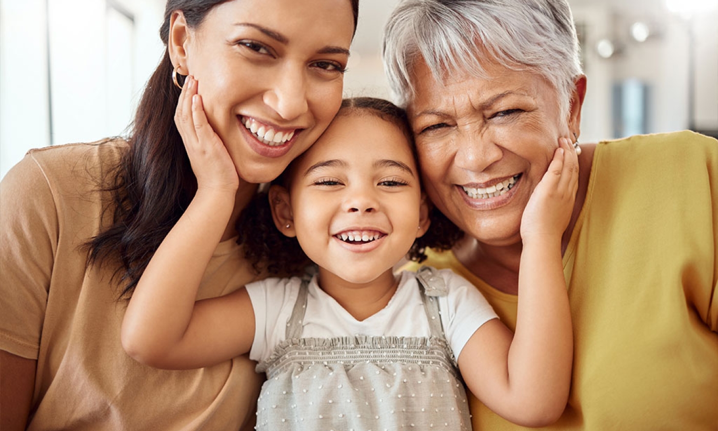 a female child sitting between an adult female and a female grandparent with her hands raised up and placed on the cheeks