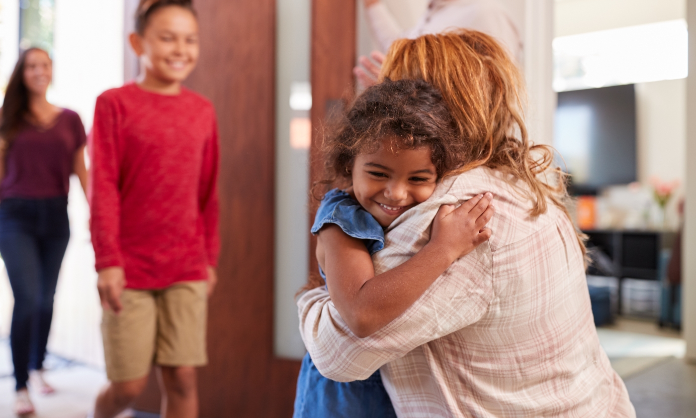 an adult woman bent over hugging an female child, an adolescent boy and adult woman behind them walking through a doorway