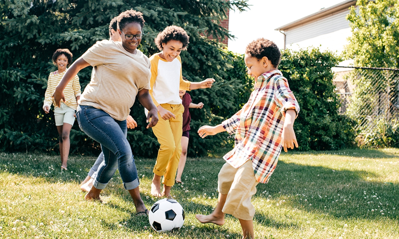 A group of adults and children playing soccer in a backyard on a sunny day.
