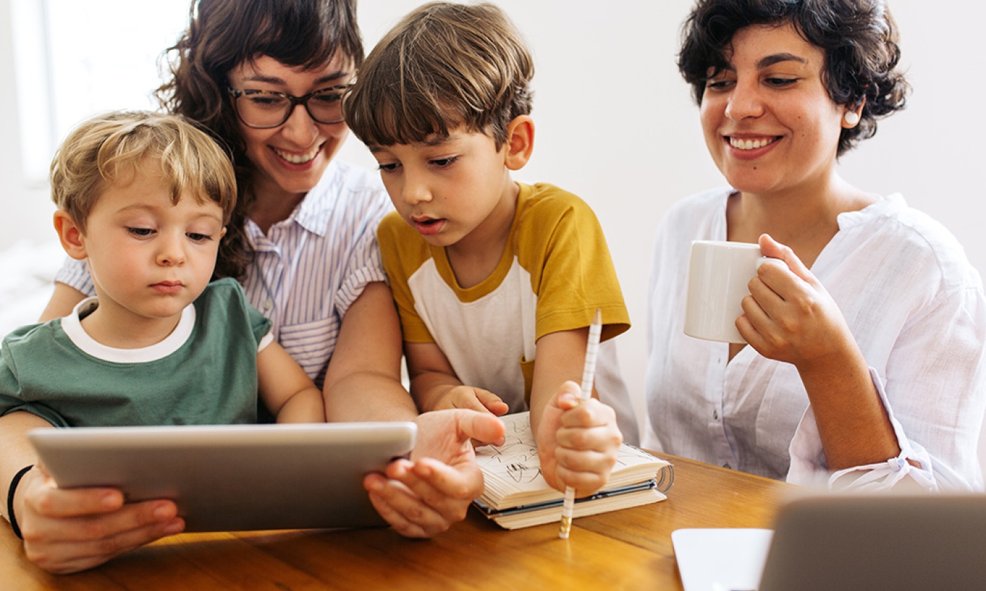 Two women and two young boys sitting together at table looking at tablet, smiling.